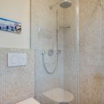 Dusche mit Glaswand, Mosaikfliesen und Duschbrause. Toilette und Waschbecken mit Flaschen.