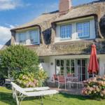 Haus mit Strohdach, Terrasse mit Tisch und Stühlen, Liegestühle auf Rasen, Blumenbeete und Sonnenschirm.