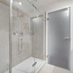 Dusche mit Glaswand und Mosaikfliesen, Nebenraum mit Schrank und Bild an der Wand.