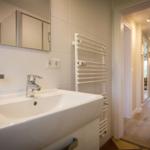 Bathroom with sink, mirror, and heater. Hallway to living area with wooden floor.