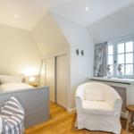 Schlafzimmer mit Bett, Sessel und Fenster. Holzboden und weiße Wände.