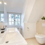 Badezimmer mit Doppelwaschbecken, Toilette und Fenster mit Blick auf Bäume.