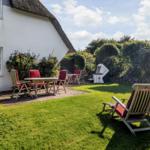 Backyard with lawn, patio, and garden chairs in front of a white house with thatched roof.