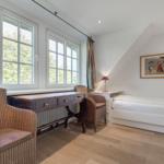 Room with desk, bed, and windows overlooking trees.