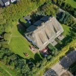 House with thatched roof, surrounded by lawn and trees. Outdoor area with red garden furniture.