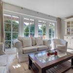 Living room with sofa, armchairs, and glass-top table. Large windows and white cabinets.