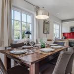 Dining room with table, chairs, and windows. Kitchen with red backsplash and white cabinets.