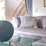 Ein Sofa mit Kissen und Decke vor einem Glas-Tisch. Hintergrund: Treppe und Fenster.