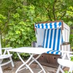 Behind green bushes, a white terrace with a blue-and-white striped beach chair and white chairs.