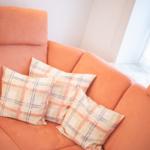 Orangefarbener Sofa mit drei karierten Kissen und Fenster im Hintergrund.