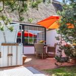 Terrasse mit orangefarbenem Sonnenschirm und weißem Liegestuhl vor einem Haus mit Strohdach.