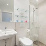 Badezimmer mit Dusche, Waschbecken und Toilette. Glasregale und Spiegel sichtbar.