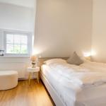 Schlafzimmer mit Bett, Nachttisch und Fenster. Holzboden und Wandheizung.