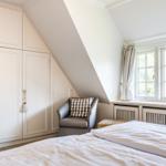 Schlafzimmer mit Dachschrägen, weißem Schrank und Fenster mit Blick auf Bäume.