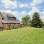Haus mit Strohdach, Rasenfläche und Blumenbeete. Gartenmöbel und Nadelbaum sind sichtbar.