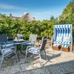 Terrasse mit Tisch, Stühlen und Strandkorb. Hintergrund: Grünflächen und Dachhäuser.