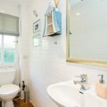 Badezimmer mit Toilette, Waschbecken und Spiegel. Fenster mit Rollladen und Wandregal.
