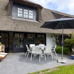 Terrasse mit Tisch, Stühlen und Sonnenschirm vor einem Haus mit Strohdach.