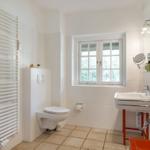 Badezimmer mit Toilette, Waschbecken und Fenster. Weiße Fliesen und Holzboden.