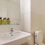 Badezimmer mit Waschbecken, Spiegel und Toilette. Auf dem Regal stehen grüne Flaschen.