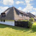 Haus mit strohgedecktem Dach und weißer Fassade, umgeben von Grünfläche und Hecken.