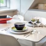 Tisch mit Geschirr und Gläsern, Fenster und Küchenbereich im Hintergrund.