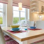 Dining area with wooden table, chairs, and windows. Kitchen in background.