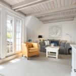 Wohnzimmer mit Sofa, Sessel und großen Fenstern. Decke mit Holzbalken und Wandbild.