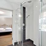Shower with glass enclosure and view into bedroom with bed.