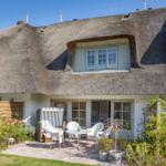Haus mit Strohdach, Terrasse mit Tisch und Stühlen, Gartenbereich mit Pflanzen.
