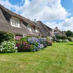 Haus mit Strohdach, Blumenbeete und Rasenfläche unter blauem Himmel.