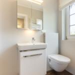 Badezimmer mit Waschbecken, Toilette und Dusche. Fenster mit Pflanze.