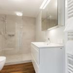 Badezimmer mit Dusche, Waschbecken und Heizung. Holzboden und weiße Fliesen.