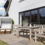 Terrasse mit Holztisch, Bänken und Strandkorb. Hintergrund: weißer Hauswand mit Fenstern.