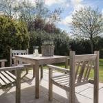 Holztisch mit Stühlen auf Terrasse, Pflanze und Kerze im Hintergrund.