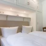 Schlafzimmer mit Bett, weißen Kissen und Holzstuhl. Oberhalb des Bettes befinden sich Schränke und eine Ablage.