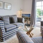 Living room with sofa, armchairs, glass table, and TV. View to balcony with garden.