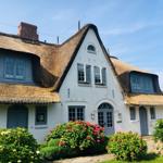 Haus mit Strohdach, weißen Wänden und blauen Fenstern, umgeben von Blumenbeeten.