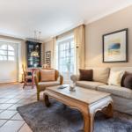 Living room with sofa, coffee table, and armchair. Windows, wall art, and kitchen area visible.