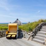 Ein gelber Strandkorb steht auf einer Holzterrasse mit Steintreppe und Blick auf das Meer.
