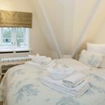 Bedroom with sloped ceiling, bed with blue and white bedding, and window with gold roman shades.