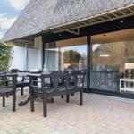 Terrasse mit Tisch und Stühlen unter strohgedecktem Dach. Große Glasfront zeigt Innenraum.