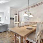 Dining room with wooden table, white chairs, and decorative tiled walls. Living area with sofa and window.