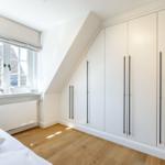 Schlafzimmer mit Dachschrägen, weißem Schrank und Holzboden. Fenster mit Blick auf Dächer.