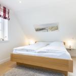 Schlafzimmer mit Doppelbett, Holzboden und zwei Fenstern mit roten Gardinen.