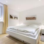 Schlafzimmer mit weißem Bett, Holzboden und Fenster mit gelben Vorhängen.