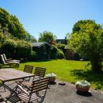 Garten mit Terrasse, Holztisch, Stühlen und Blumenkübeln. Grasfläche und Bäume im Hintergrund.
