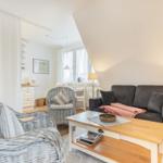 Wohnzimmer mit grauem Sofa, weißen Rattan-Stühlen und Holztisch. Dachschrägen und Fenster sichtbar.