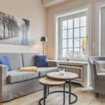 Ein Wohnzimmer mit Sofa, Sessel und Tisch. An der Wand hängt ein Bild. Durch Fenster ist die Aussicht aufs Meer.