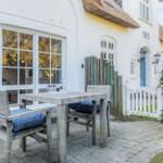 Wooden table with chairs on paved courtyard in front of white house with thatched roof.